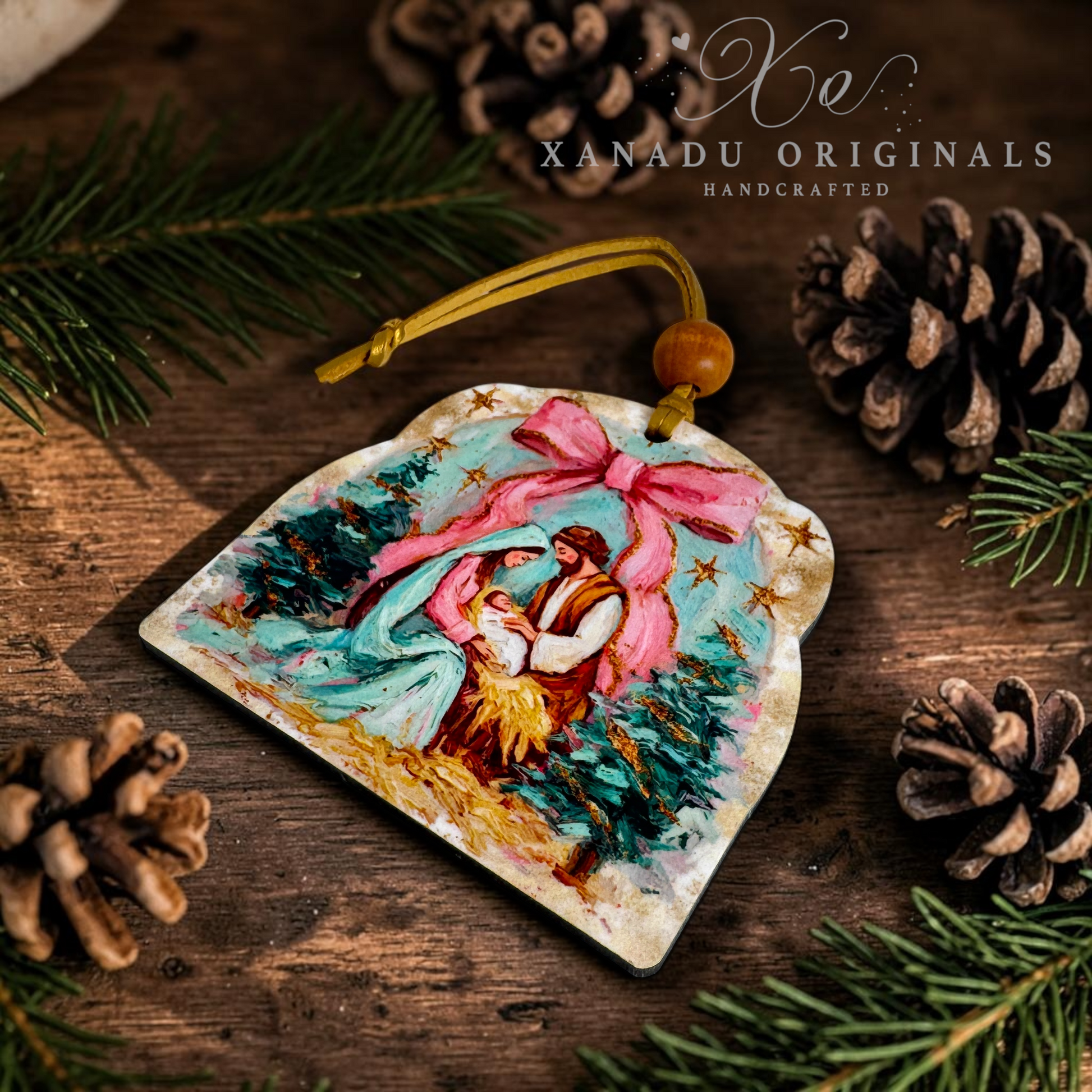 A handcrafted wooden Christmas ornament featuring an illustrated nativity scene with a pink bow, resting on a rustic wooden surface surrounded by pine needles and pinecones. The brand name 'XANADU ORIGINALS' is visible in the upper right corner.