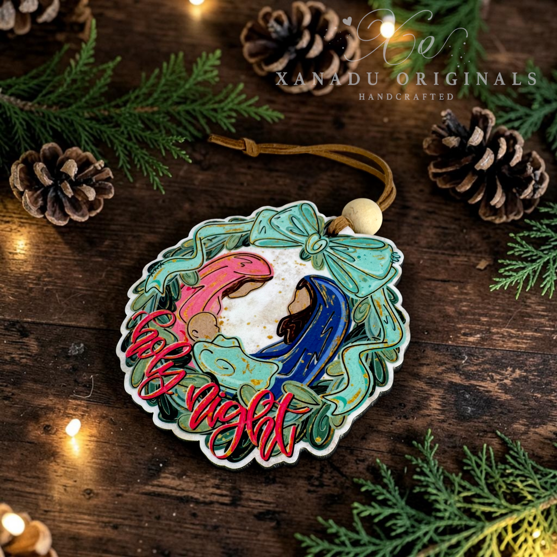A handcrafted Christmas ornament featuring a nativity scene with Mary, Joseph, baby Jesus, and an angel in colorful pastel tones, hanging from a brown cord on a rustic wooden surface surrounded by pine branches, pinecones, and warm fairy lights. The brand name 'XANADU ORIGINALS' is visible in the upper right.
