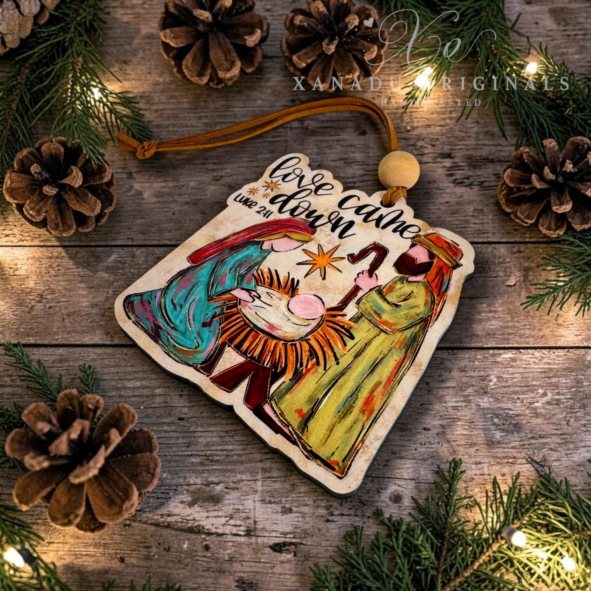 A hand-painted wooden Christmas ornament featuring Mary, Joseph, and baby Jesus with the text 'Love came down', displayed on a rustic wooden surface decorated with pine branches, pinecones, and warm fairy lights. A watermark for 'Xanadu Originals' is visible in the upper right.