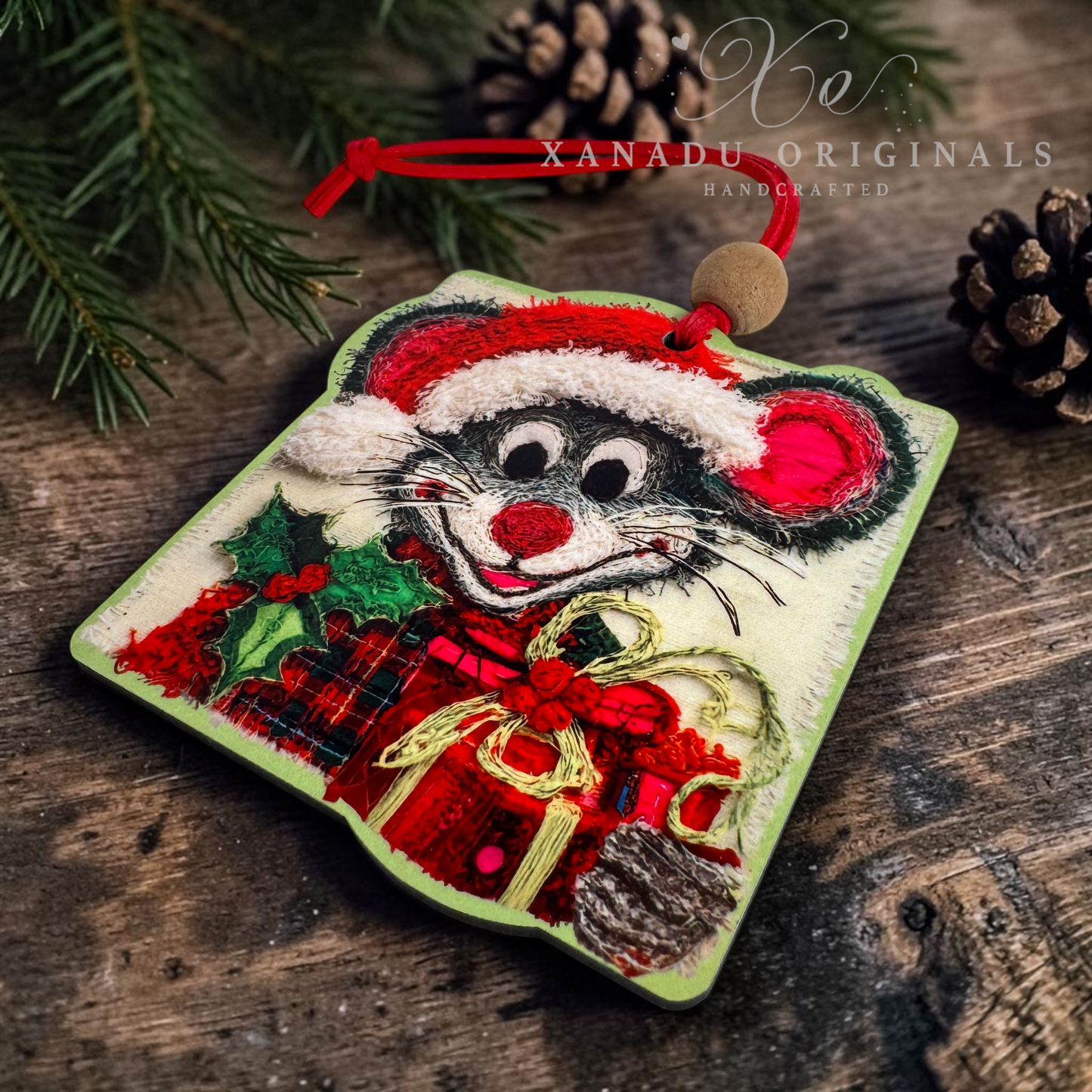 A handcrafted embroidered Christmas ornament shaped like a mouse wearing a Santa hat and holding red wrapped gifts, displayed on a rustic wooden surface with pine branches and a pinecone. The watermark 'XANADU ORIGINALS HANDCRAFTED' is visible in the upper right.