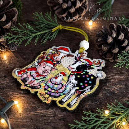 A wooden Christmas ornament featuring a festive illustration of a pig, goose, chicken, and cow wearing Santa hats and holiday decorations, hanging on a dark wooden surface with pine branches, pinecones, and warm string lights. The brand name 'XANADU ORIGINALS' is watermarked at the top.