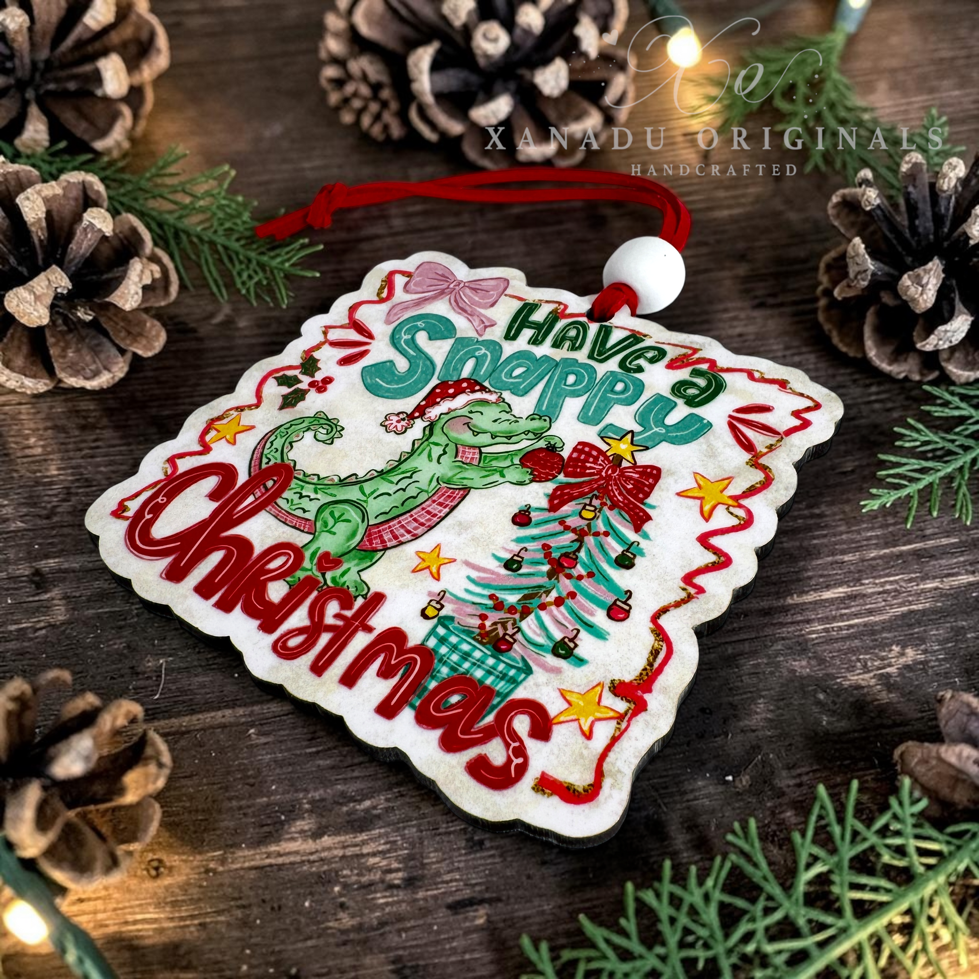 A square white embroidered Christmas ornament with scalloped edges, featuring a green alligator, a decorated Christmas tree, red lettering that reads 'Have a Snappy Christmas,' and a red hanging cord. The ornament is photographed on a rustic wooden surface surrounded by pinecones and evergreen branches.