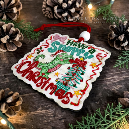 A square white embroidered Christmas ornament with scalloped edges, featuring a green alligator, a decorated Christmas tree, red lettering that reads 'Have a Snappy Christmas,' and a red hanging cord. The ornament is photographed on a rustic wooden surface surrounded by pinecones and evergreen branches.