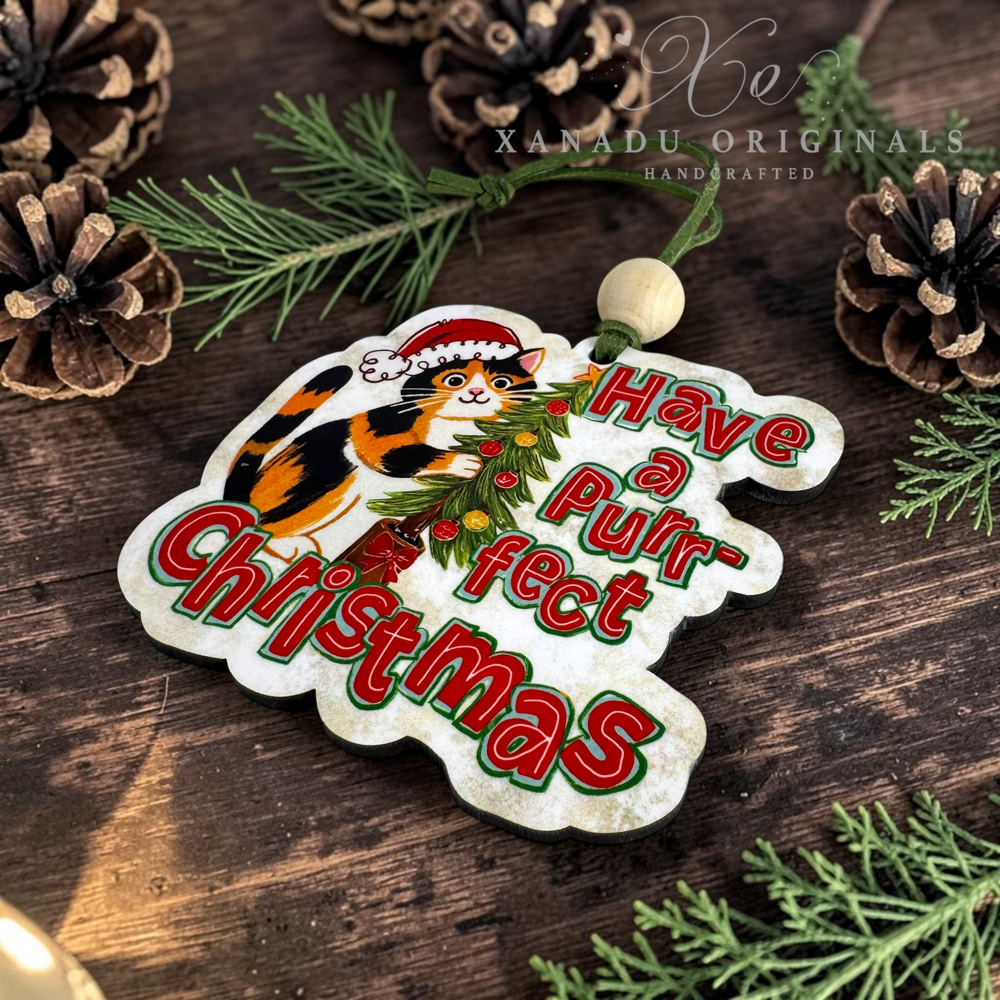 A decorative Christmas ornament shaped like a cat silhouette with the phrase “Have a Purrfect Christmas” in red and green lettering, featuring an illustrated calico cat wearing a Santa hat. The ornament hangs from a green cord with a wooden bead and is styled on a dark wood surface with pine branches and pinecones.