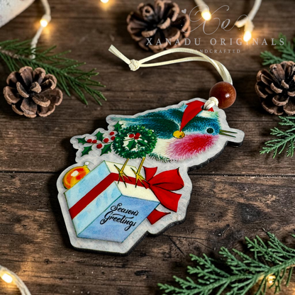 A handmade Christmas ornament shaped like a robin holding a wrapped gift, decorated with a green wreath and red hat, hanging on a rustic wooden surface with pine branches, pinecones, and warm string lights. The ornament has a white hanging loop with a wooden bead and the brand name 'XANADU ORIGINALS' is watermarked at the top.