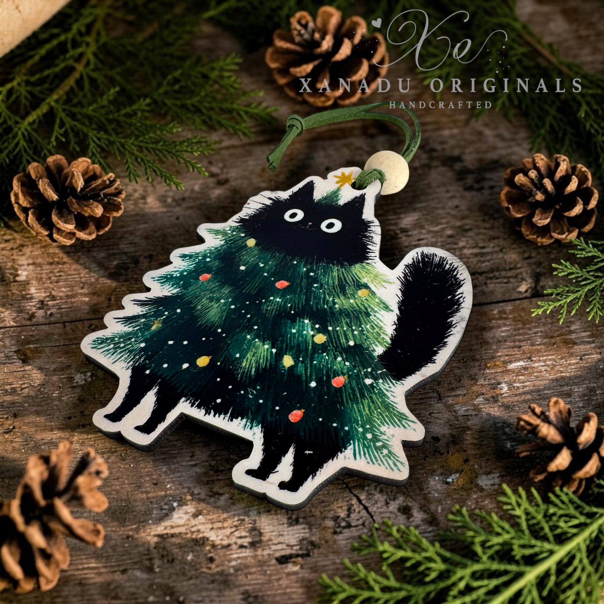 A Christmas ornament shaped like a black cat wearing a green Christmas tree costume, decorated with colorful ornaments and a small gold star, hanging from a green cord. The ornament is displayed on a rustic wooden surface surrounded by pine needles, pinecones, and evergreen branches.