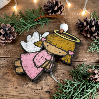 A handcrafted wooden angel ornament with a pink dress, white wings, and a gold halo, decorated with a brown cord and white bead, resting on a rustic wooden surface with pinecones and evergreen branches.