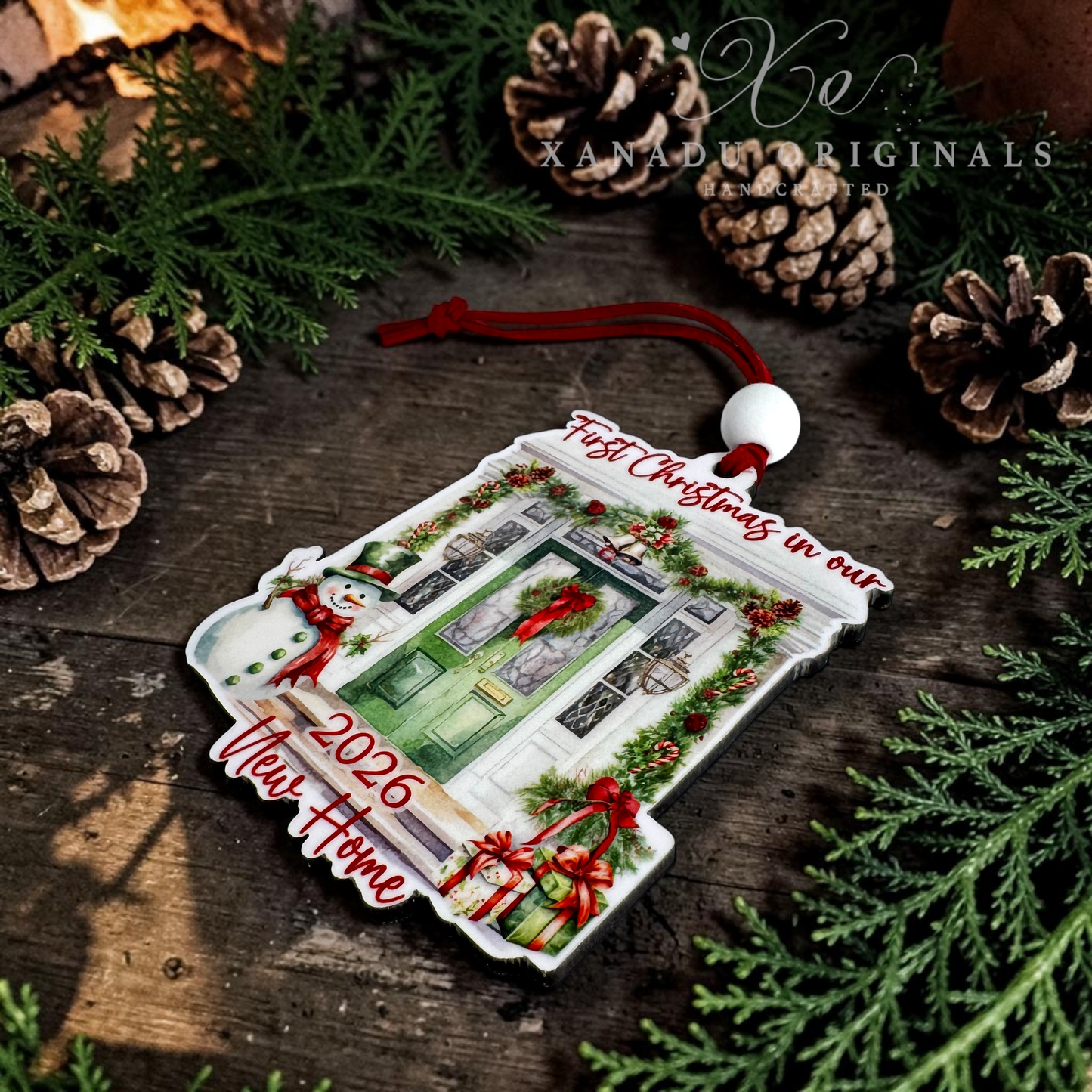 A custom Christmas ornament shaped like a front door, decorated with a green door, red bows, holly, and snowman figures, with text reading 'First Christmas in our 2026 New Home' and 'Past Christmas in our 2025 New Home.' It is displayed on a rustic wooden surface with pine branches, pinecones, and a red hanging cord.