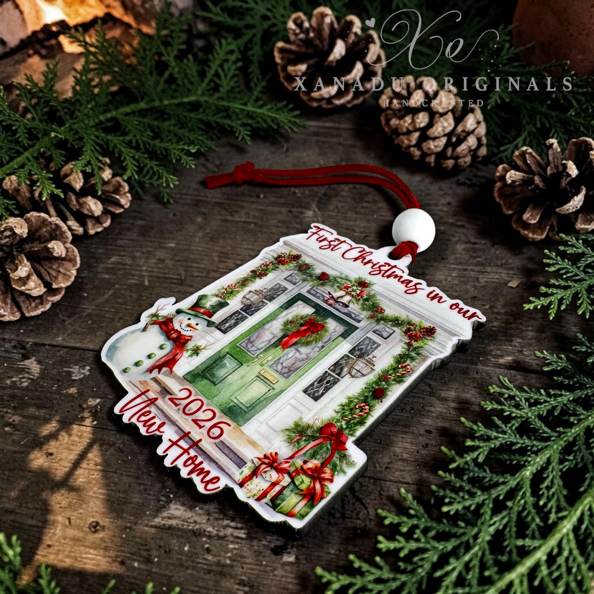A custom Christmas ornament shaped like a front door, decorated with a green door, red bows, holly, and snowman figures, with text reading 'First Christmas in our 2026 New Home' and 'Past Christmas in our 2025 New Home.' It is displayed on a rustic wooden surface with pine branches, pinecones, and a red hanging cord.