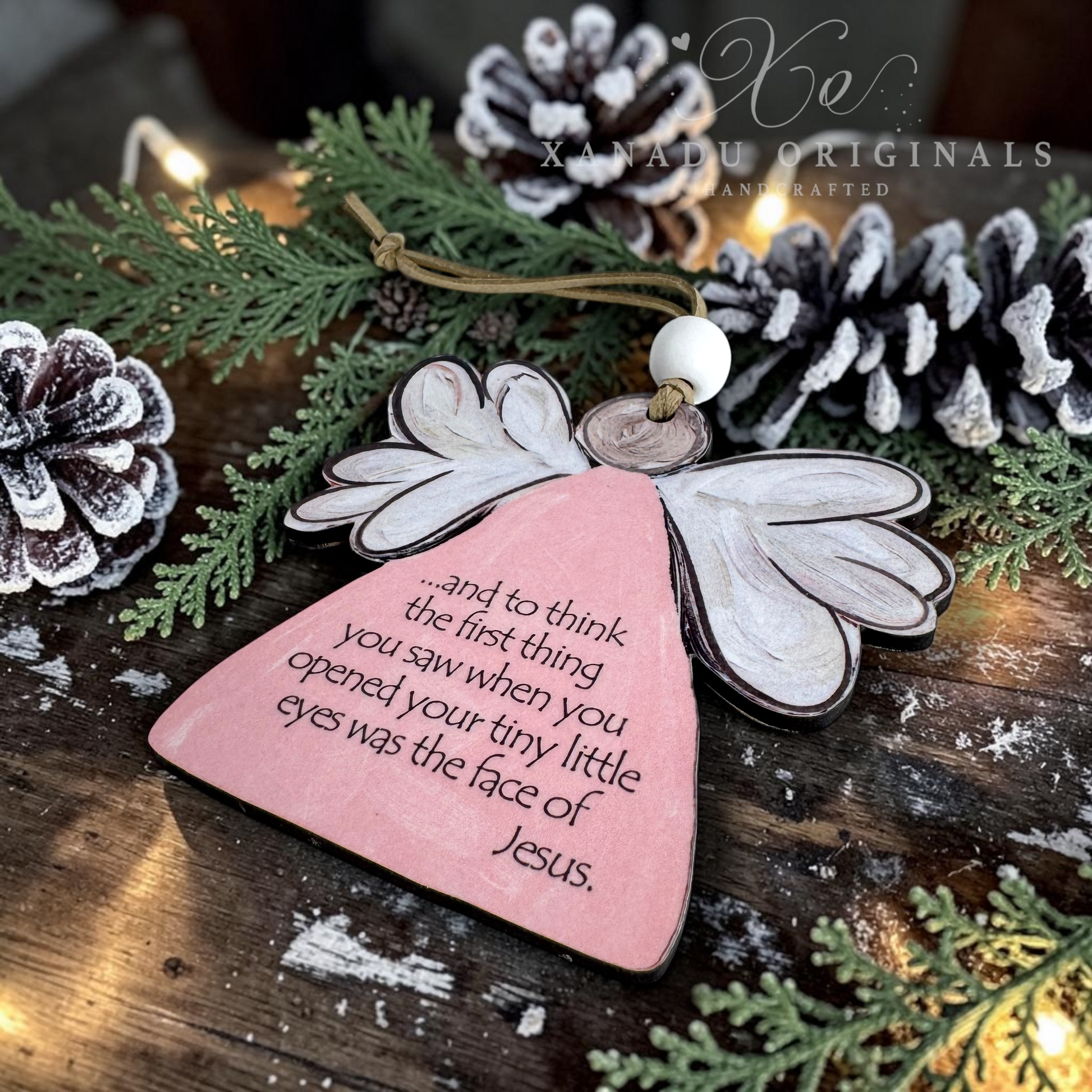 A wooden angel miscarriage ornament with a pink body, white wings, and a printed Bible verse, displayed on a rustic wooden surface with pine branches, snow-dusted pinecones, and warm string lights. A watermark for 'XANADU ORIGINALS' is visible in the upper right.