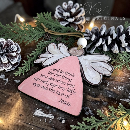 A wooden angel miscarriage ornament with a pink body, white wings, and a printed Bible verse, displayed on a rustic wooden surface with pine branches, snow-dusted pinecones, and warm string lights. A watermark for 'XANADU ORIGINALS' is visible in the upper right.
