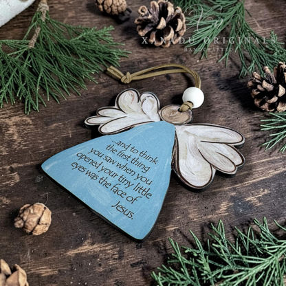 A wooden miscarriage angel ornament with white wings, a blue triangular body, and a printed inspirational quote, displayed on a dark wooden surface with pine branches and pinecones.