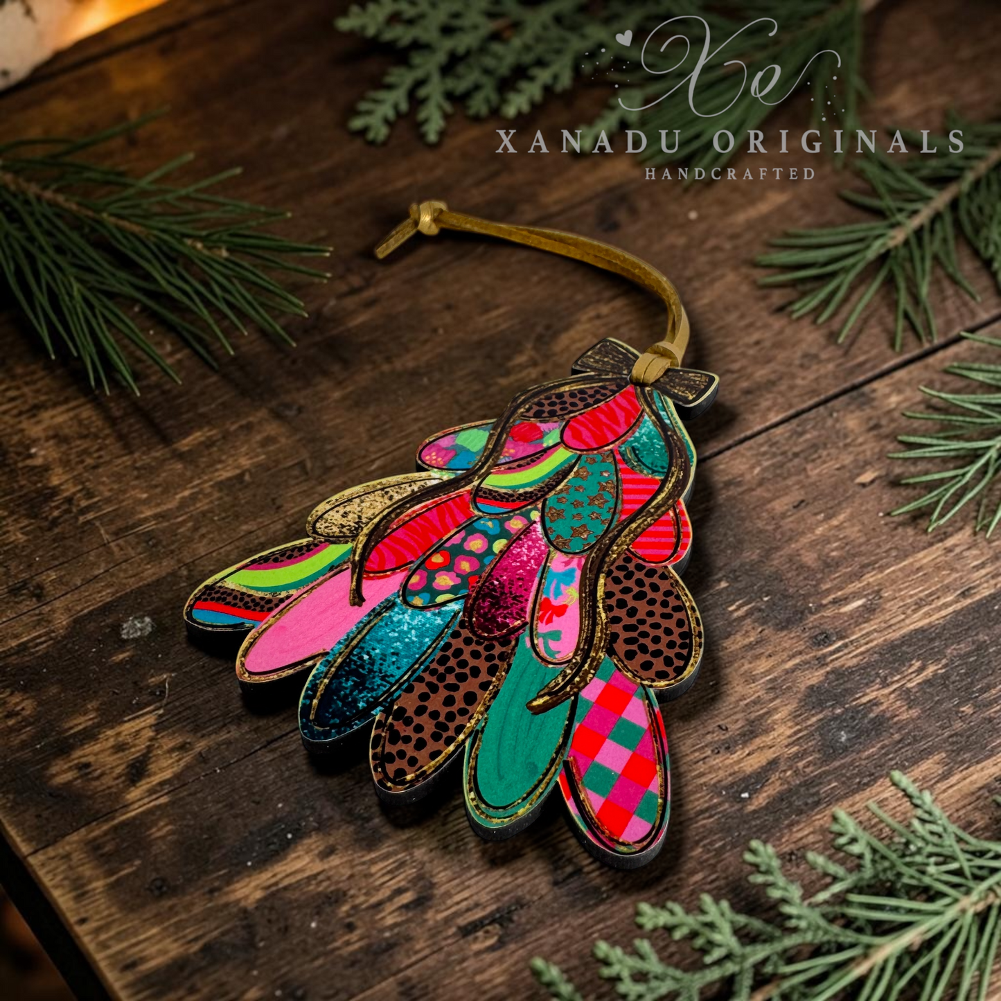 A colorful handcrafted leather ornament shaped like a stylized bird or feather cluster, laid on a rustic dark wood surface with pine branches around it. The ornament features multiple layered feather-like shapes in bright pink, teal, red, green, brown, and glittery finishes, with a tan hanging cord.