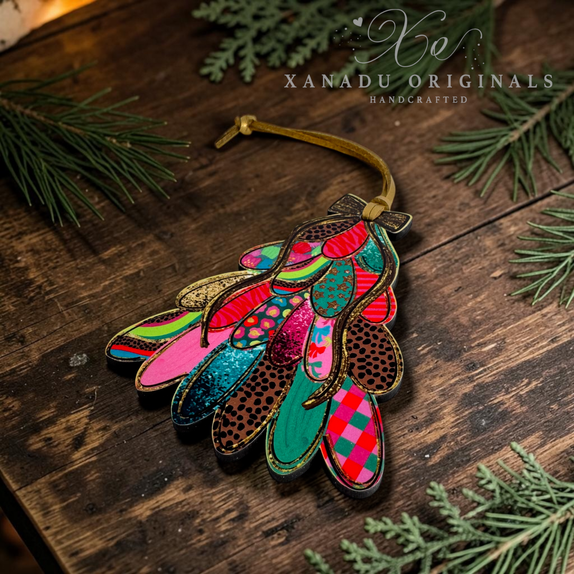 A colorful handcrafted leather ornament shaped like a stylized bird or feather cluster, laid on a rustic dark wood surface with pine branches around it. The ornament features multiple layered feather-like shapes in bright pink, teal, red, green, brown, and glittery finishes, with a tan hanging cord.
