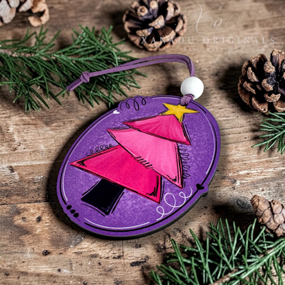 A purple oval ornament featuring a stylized pink Christmas tree with a yellow star, photographed on a rustic wooden surface with pine branches and pinecones around it. A white bead and purple loop are attached at the top.