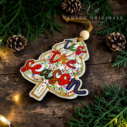 A handcrafted Christmas ornament shaped like a tree with the phrase 'Tis the season' in colorful icing, displayed on a rustic wooden surface with pine branches, pinecones, and warm string lights. The brand name 'XANADU ORIGINALS' is visible as a watermark.