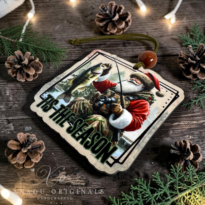 A Christmas ornament featuring a festive illustration of Santa Claus fishing with the text 'TIS THE SEASON' in large green letters. The ornament is displayed on a rustic dark wood background surrounded by pine branches, pinecones, and warm string lights.