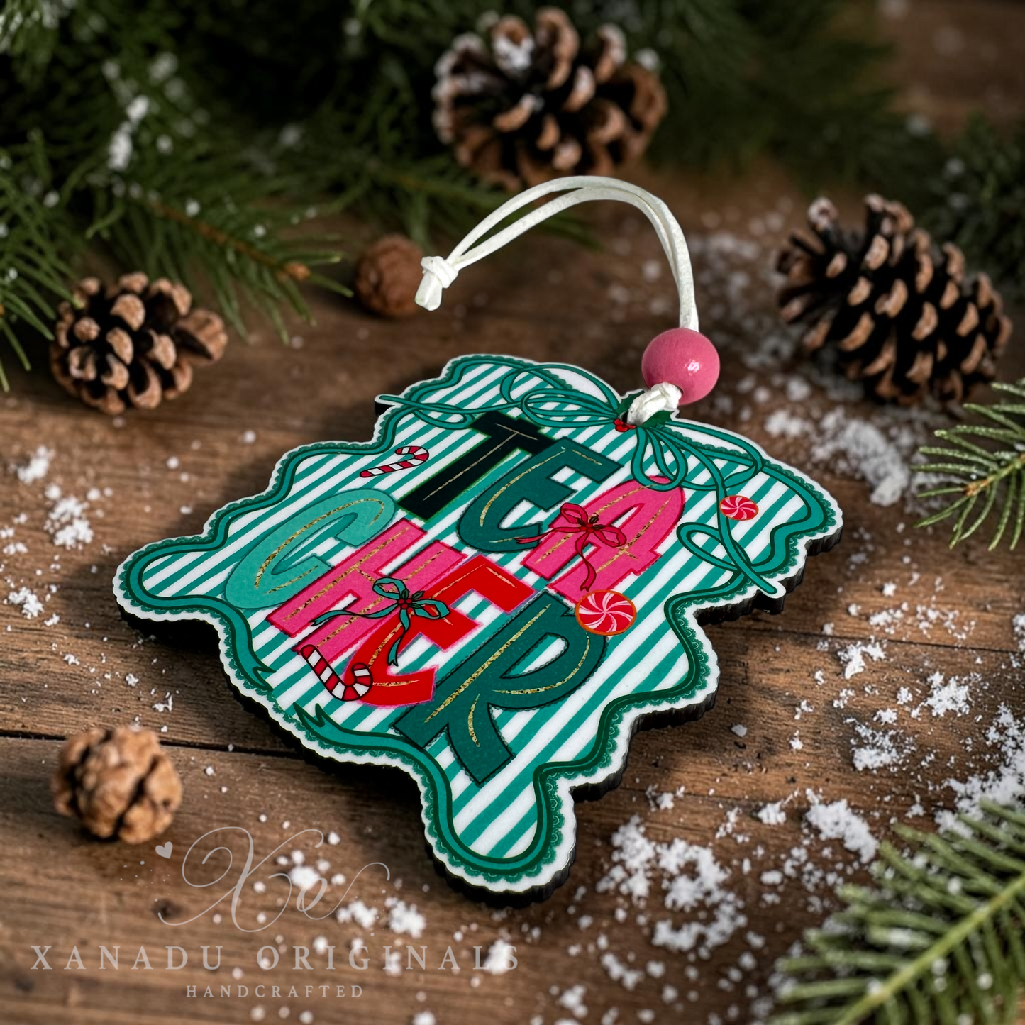 A handcrafted Christmas ornament shaped like a snowflake, featuring a green and white striped background with the word 'TEACHER' in large green letters, wrapped presents, and candy canes. The ornament is displayed on a rustic wooden surface with pine branches, pinecones, and white faux snow, with a white hanging string and pink bead.