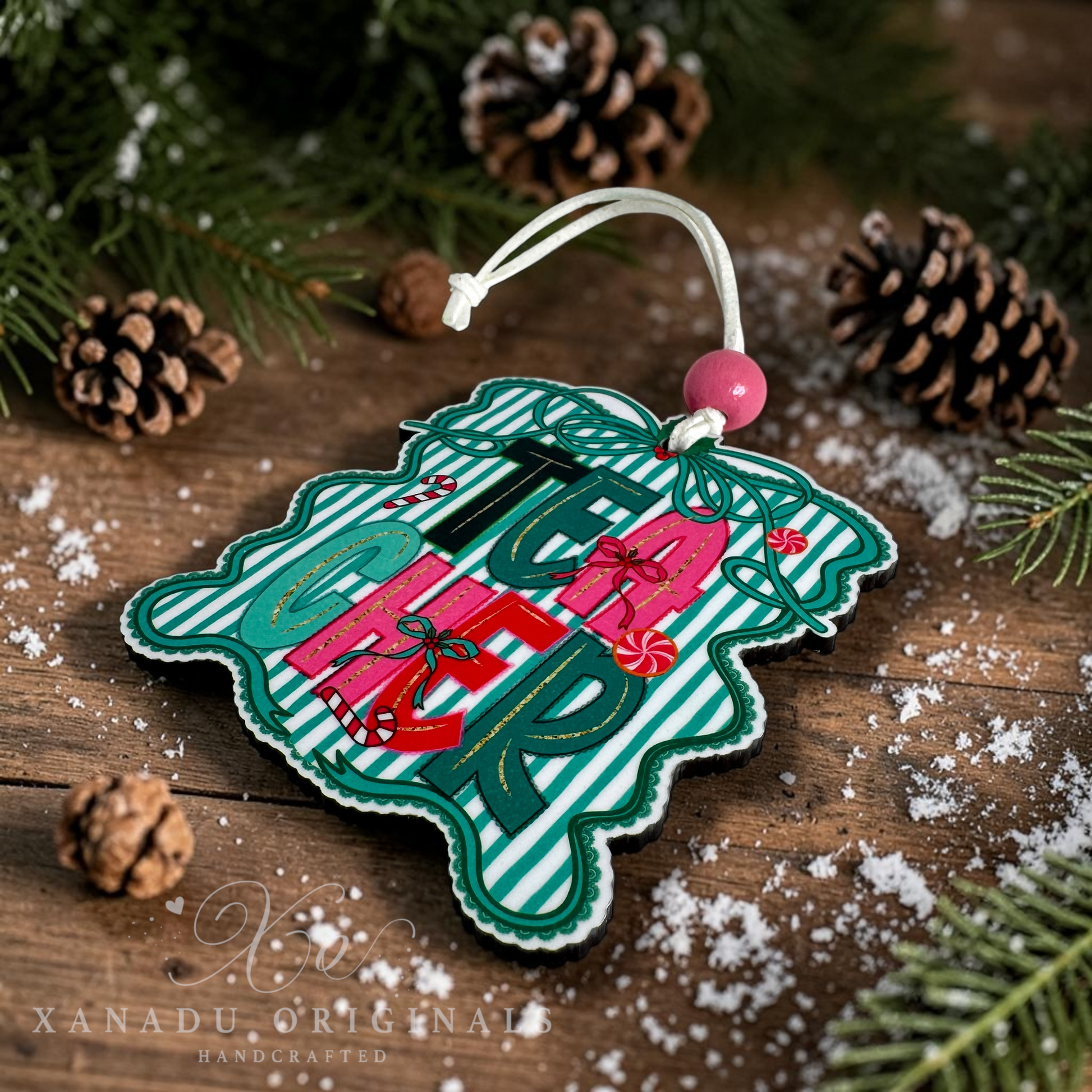A handcrafted Christmas ornament shaped like a snowflake, featuring a green and white striped background with the word 'TEACHER' in large green letters, wrapped presents, and candy canes. The ornament is displayed on a rustic wooden surface with pine branches, pinecones, and white faux snow, with a white hanging string and pink bead.