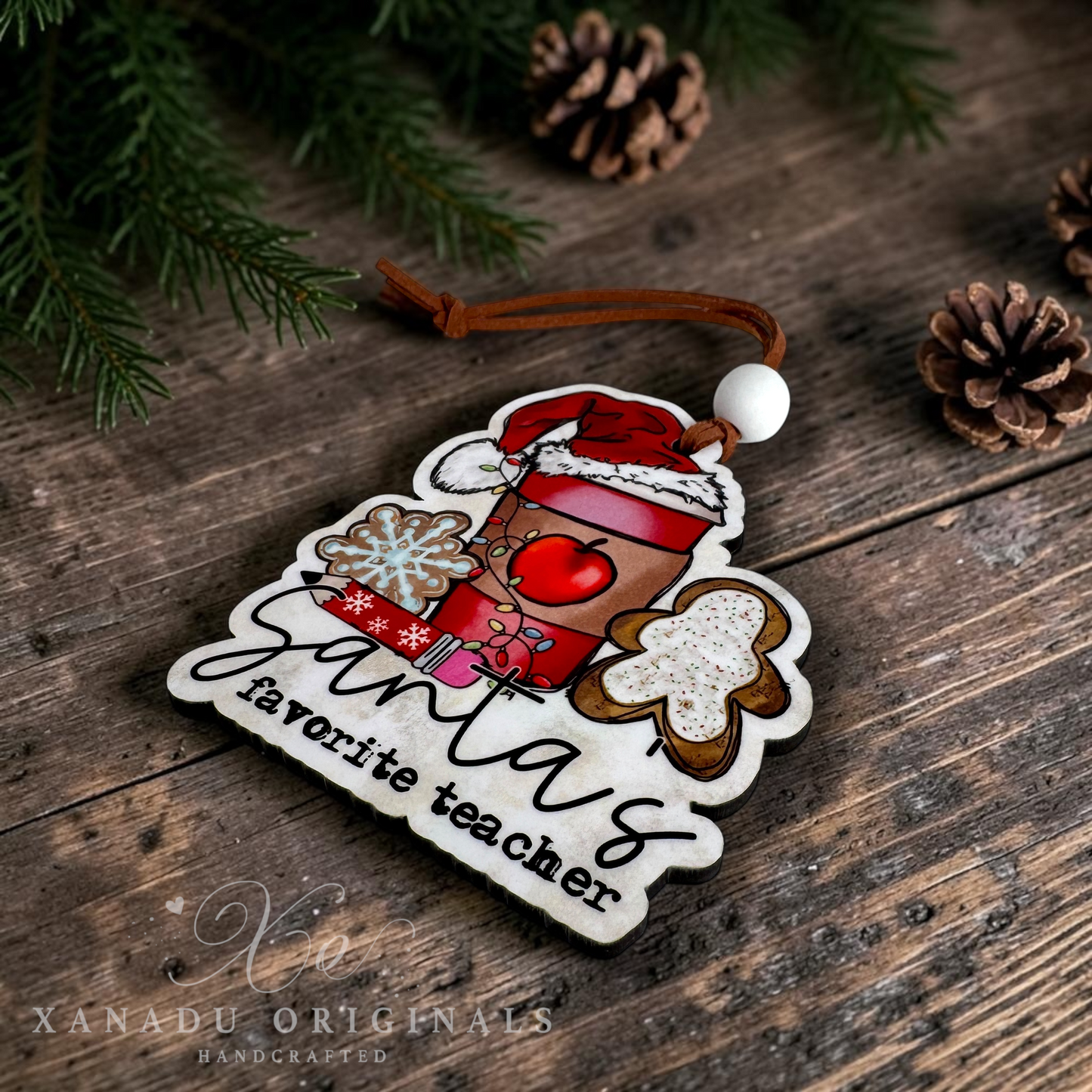 A handcrafted wooden Christmas ornament with a die-cut shape, featuring a red coffee cup with a Santa hat, gingerbread cookies, a snowflake, and the text 'Santa's favorite teacher'. The ornament is displayed on a rustic wooden surface with pine branches and pinecones.