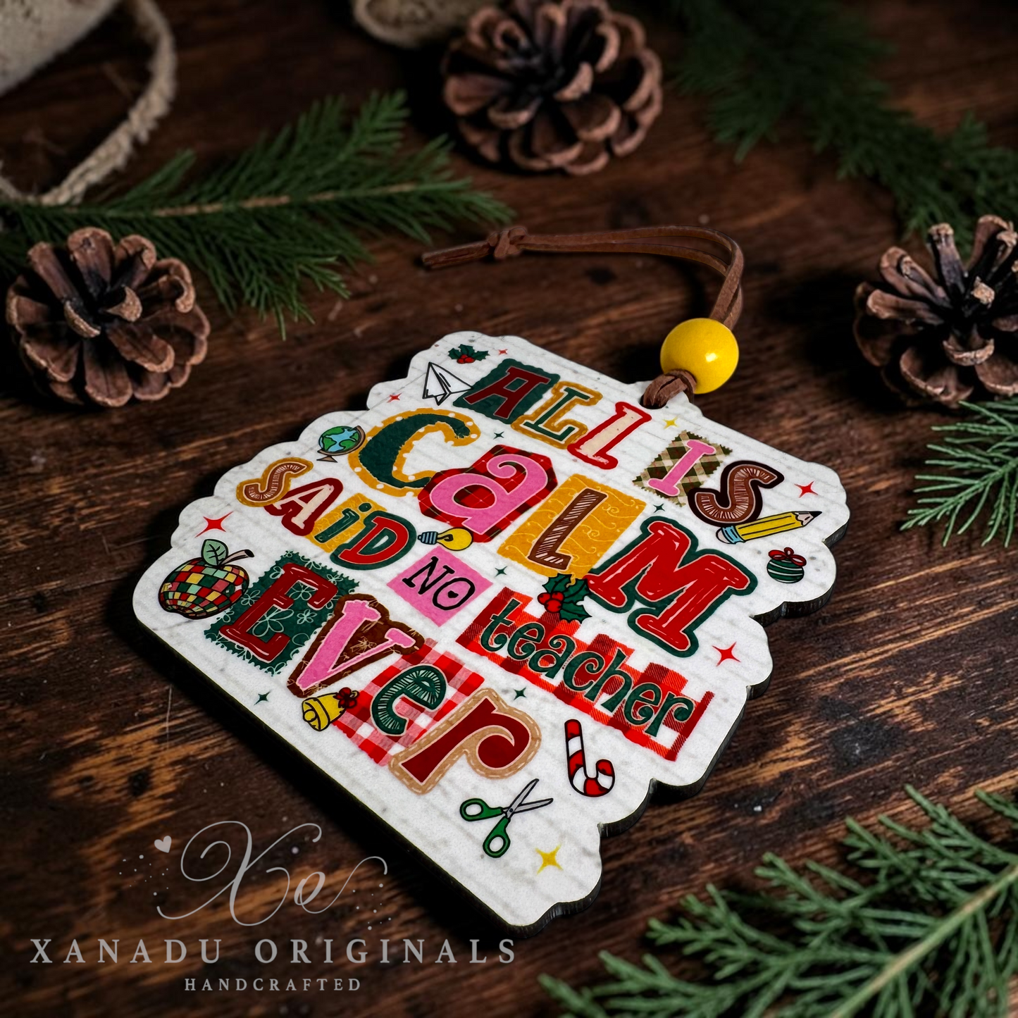 A handcrafted Christmas ornament with the phrase 'ALL IS CALM NO TEACHER EVER' in colorful retro lettering, displayed on a rustic wooden surface with pinecones and evergreen branches. The ornament has a white scalloped edge and is attached to a brown cord with a yellow bead.