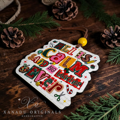 A handcrafted Christmas ornament with the phrase 'ALL IS CALM NO TEACHER EVER' in colorful retro lettering, displayed on a rustic wooden surface with pinecones and evergreen branches. The ornament has a white scalloped edge and is attached to a brown cord with a yellow bead.
