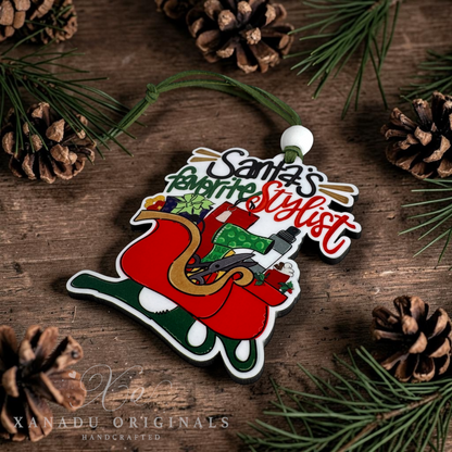 A handcrafted wooden Christmas ornament shaped like Santa's sleigh, featuring the text 'Santa's Favorite Stylist' in stylized lettering. The ornament is decorated with red, green, gold, and white details and hangs from a green cord on a rustic wooden surface surrounded by pine needles and pinecones.