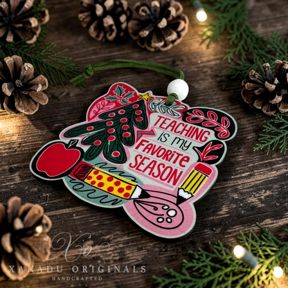 A handcrafted wooden Christmas ornament shaped like a school desk, featuring the text 'TEACHING IS MY FAVORITE SEASON' in red, with illustrations of a Christmas tree, apple, pencil, and paintbrush. The ornament is displayed on a rustic wooden surface surrounded by pinecones, evergreen branches, and warm string lights.