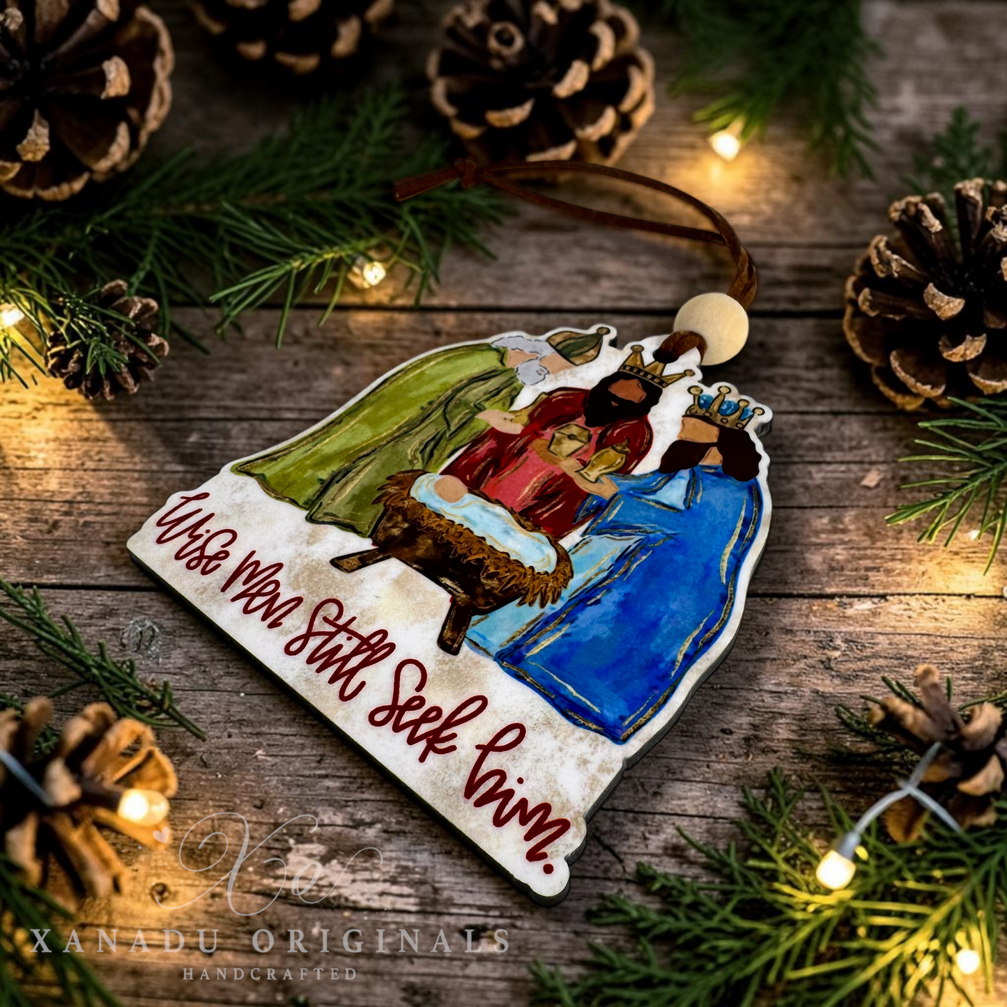 A handcrafted wooden Christmas ornament featuring a nativity scene with Mary, Joseph, baby Jesus in a manger, and an angel, along with the hand-lettered text 'Wise Men Still Seek Him.' The ornament is displayed on a rustic wooden surface surrounded by pine branches, pinecones, and warm fairy lights.