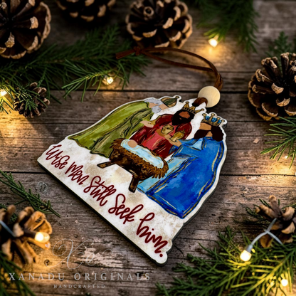 A handcrafted wooden Christmas ornament featuring a nativity scene with Mary, Joseph, baby Jesus in a manger, and an angel, along with the hand-lettered text 'Wise Men Still Seek Him.' The ornament is displayed on a rustic wooden surface surrounded by pine branches, pinecones, and warm fairy lights.