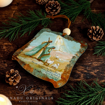 A handcrafted Christmas ornament featuring a painted nativity scene with the Wise men and angel, and sheep, hanging from a brown cord with a white bead. The ornament is displayed on a rustic dark wood surface surrounded by pine needles and pinecones, with a watermark for 'Xanadu Originals Handcrafted' at the bottom.