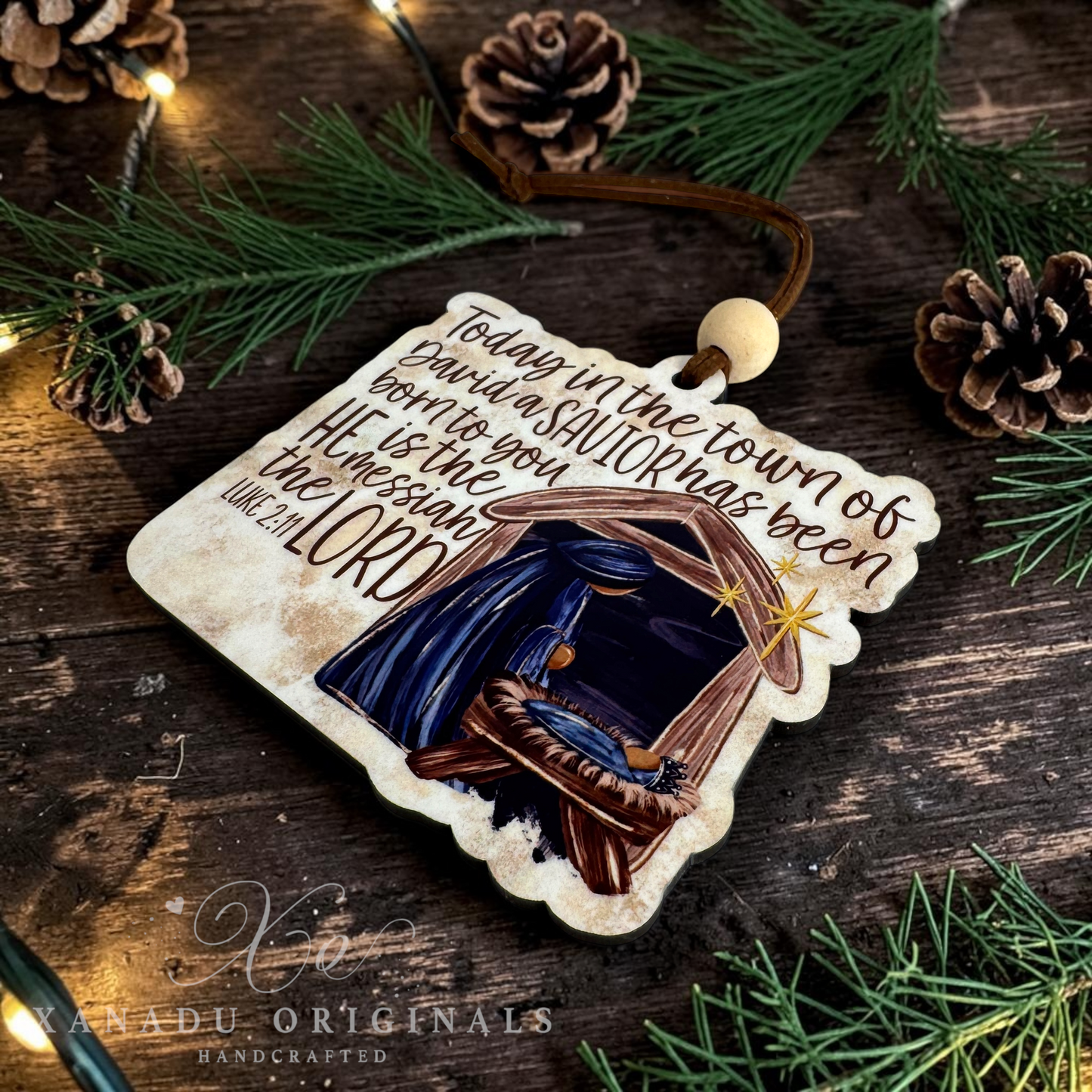 A handcrafted wooden Christmas ornament featuring a nativity scene and scripture, styled on a rustic dark wood background with pine branches, pinecones, and warm fairy lights. The ornament is branded with 'Xanadu Originals Handcrafted' at the bottom.