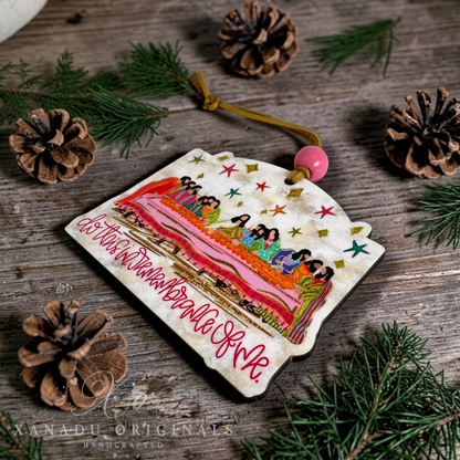 A handcrafted Christmas ornament  featuring a colorful illustration of people in a table and the text 'Do this in remembrance of me'. The ornament is decorated with a pink bead and a tan string, displayed on a rustic wooden surface surrounded by pine needles and pinecones.