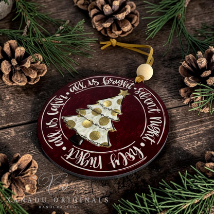 A round wooden Christmas ornament with a dark red background, white script text, and a gold-and-white patterned tree in the center. The ornament hangs from a yellow cord with a light wood bead and is photographed on a rustic dark wood surface surrounded by pine branches, pinecones, and greenery.