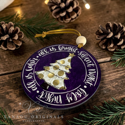 A round wooden Christmas ornament with a purple background, white script text, and a white tree with gold polka dots, hanging on a rustic wooden surface with pine branches and pinecones around it.