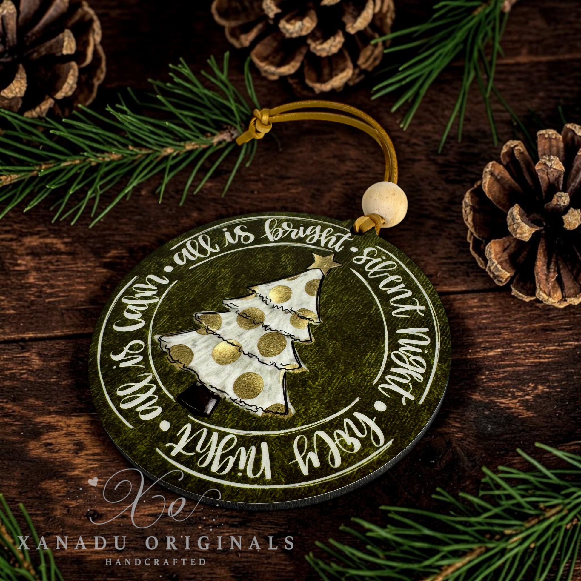 A round wooden Christmas ornament with a dark green distressed finish, featuring a 3D white and gold pine tree, the words 'all is bright and silent now', and a hanging loop. The ornament is styled on a dark wood background with pine needles and pinecones.
