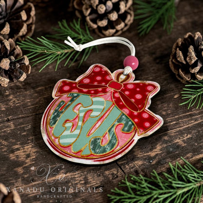 A handcrafted Christmas ornament shaped like a red bow-tied gift bag with green and pink monogrammed  letters that spell JESUS, displayed on a rustic dark wood surface with pine needles and pinecones. Xanadu Originals branding is visible in the lower-left corner.