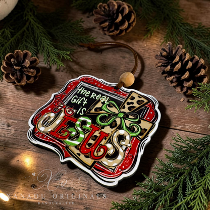 A handcrafted Christmas ornament with the text 'The Real Gift Is JESUS' in red, green, and gold lettering, styled on a rustic wooden surface with pinecones and evergreen branches.
