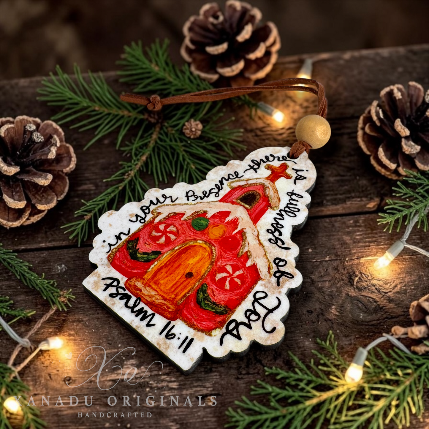 A handcrafted Christmas ornament shaped like a gingerbread church, painted with red, orange, and green details, with black handwritten scripture text. It is displayed on a rustic wooden surface surrounded by pine branches, pinecones, and warm fairy lights.