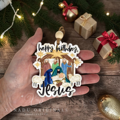 A hand holds a handcrafted wood Christmas ornament featuring a nativity scene with the text 'happy birthday, Jesus' in black script. The ornament is decorated with colorful figures, a golden star, and white sheep, shown against a rustic wooden background with festive pine branches, gift boxes, and warm string lights.
