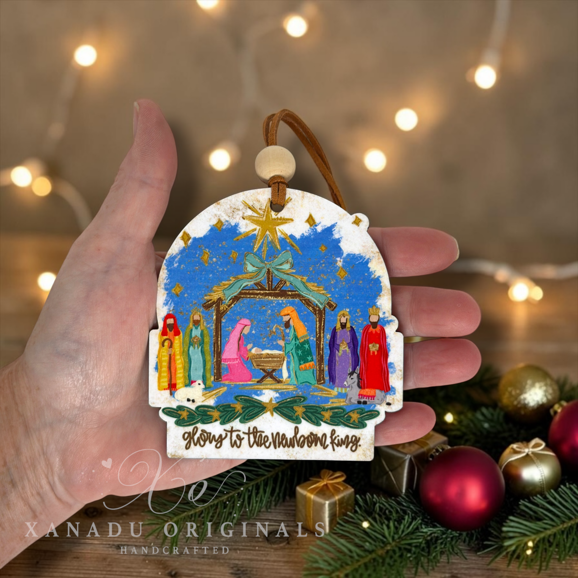 A hand holds a handcrafted Christmas ornament featuring a nativity scene with colorful figures under a blue starry sky. The ornament has a brown hanging loop and a wooden bead.