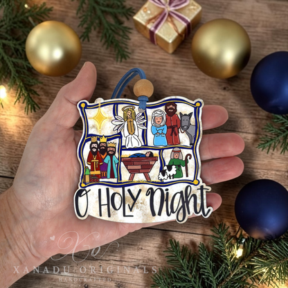 A hand holds a die-cut Christmas ornament featuring a colorful nativity scene with the text 'O Holy Night' at the bottom. The background is a rustic wooden surface decorated with gold and blue Christmas ornaments, pine branches, and a small wrapped gift box.