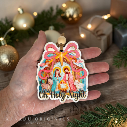 A hand holds a handcrafted Christmas ornament shaped like a Christmas tree with a colorful nativity scene and the words 'Oh Holy Night.' The background shows a cozy holiday setup with pine branches, gold ornaments, and wrapped gifts.