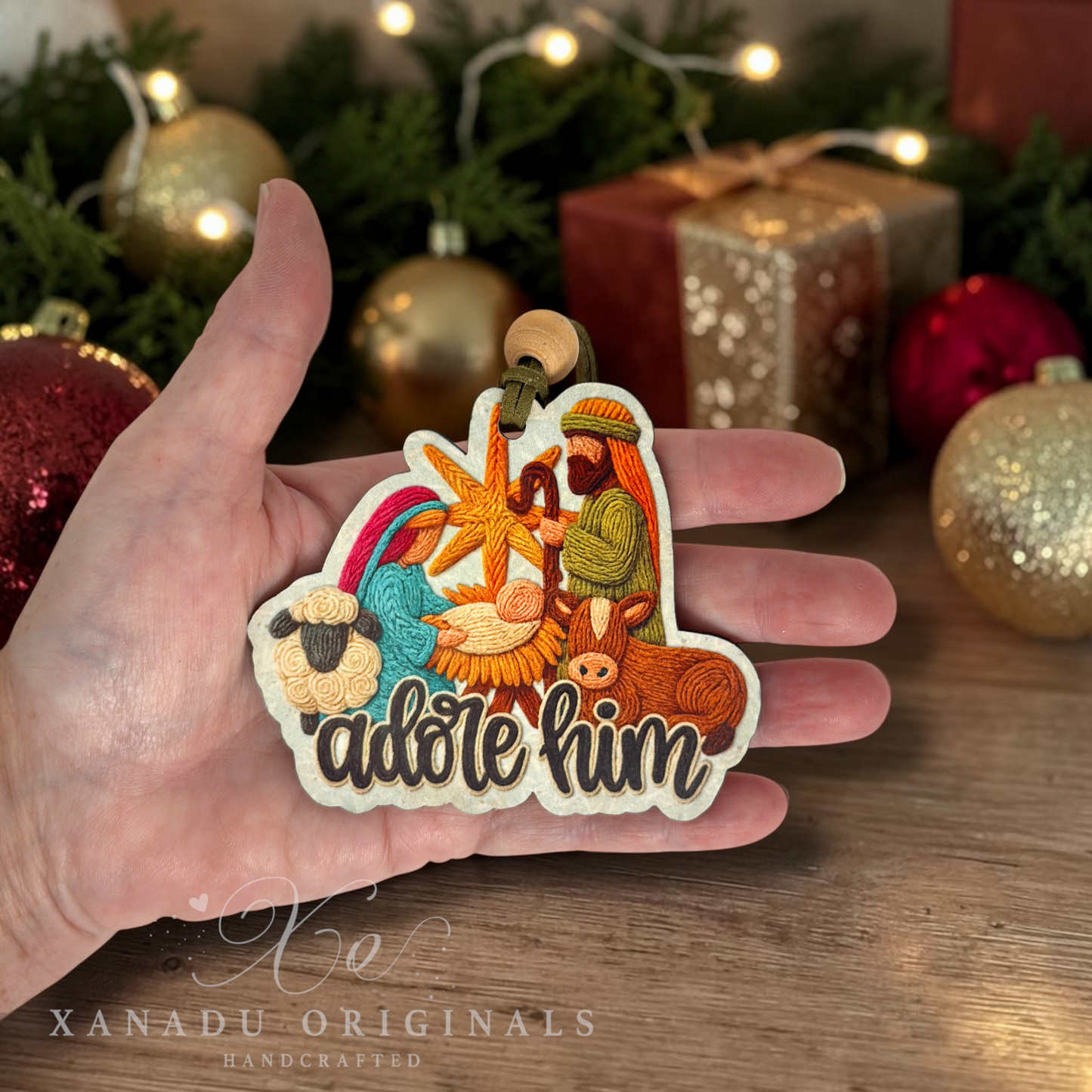 A hand holds a die-cut wooden ornament featuring an embroidered nativity scene with the text ' adore him'. The background shows a festive Christmas setting with ornaments, wrapped gifts, and warm string lights.