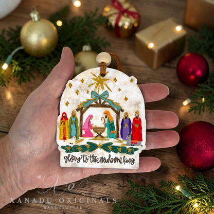 A hand holds a handcrafted Christmas ornament shaped like a rustic white sign with a Nativity scene and the text 'Glory to the Newborn King.' The background shows a wooden surface with pine branches, gold and red ornaments, wrapped gifts, and warm string lights.