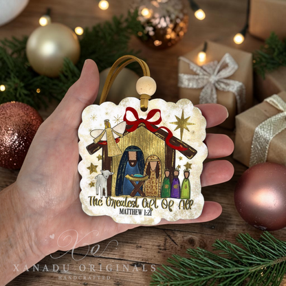 A hand holds a scalloped square Christmas ornament featuring a nativity scene with gold glitter stars, a red bow, and the text 'The Greatest Gift Of All MATTHEW 1:21'. The background shows a cozy holiday setting with wrapped gifts, pine branches, and warm lights.