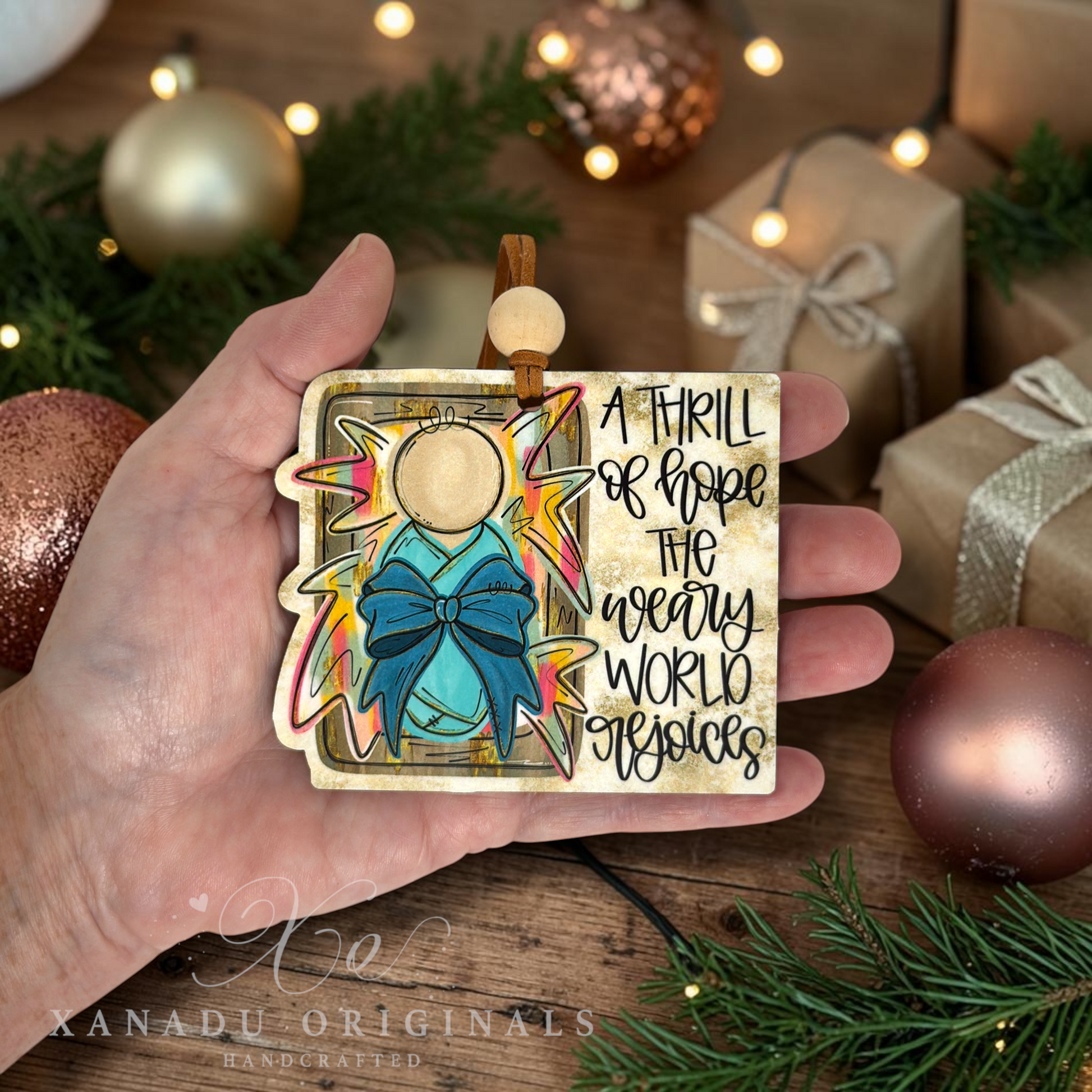 A hand holds a square wooden Christmas ornament featuring a gold angel with a blue bow, colorful starburst rays, and the text 'A THRILL OF HOPE THE WEARY WORLD OBJECTS.' The background shows a cozy holiday scene with wrapped gifts, ornaments, pine branches, and warm string lights.