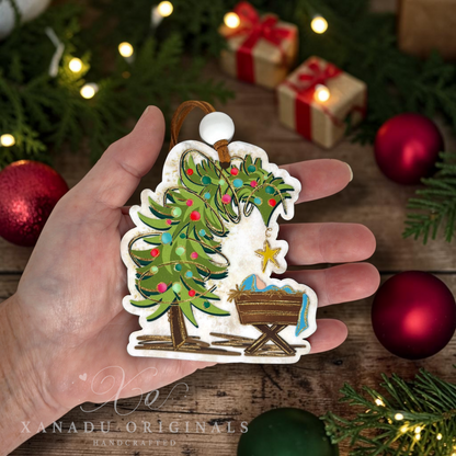 A hand holds a handcrafted Christmas ornament shaped like a decorated tree and a baby cradle, with a wooden background and festive ornaments and lights around it.