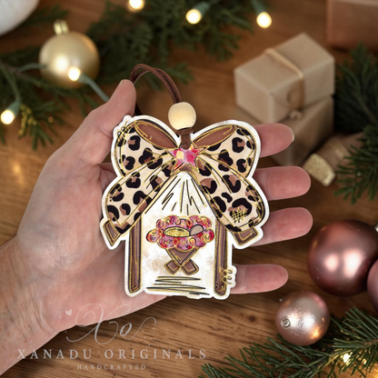 A hand holds a Christmas ornament shaped like a manger with a leopard-print bow and a pink star design, set against a cozy holiday background with pine branches, lights, and wrapped gifts.