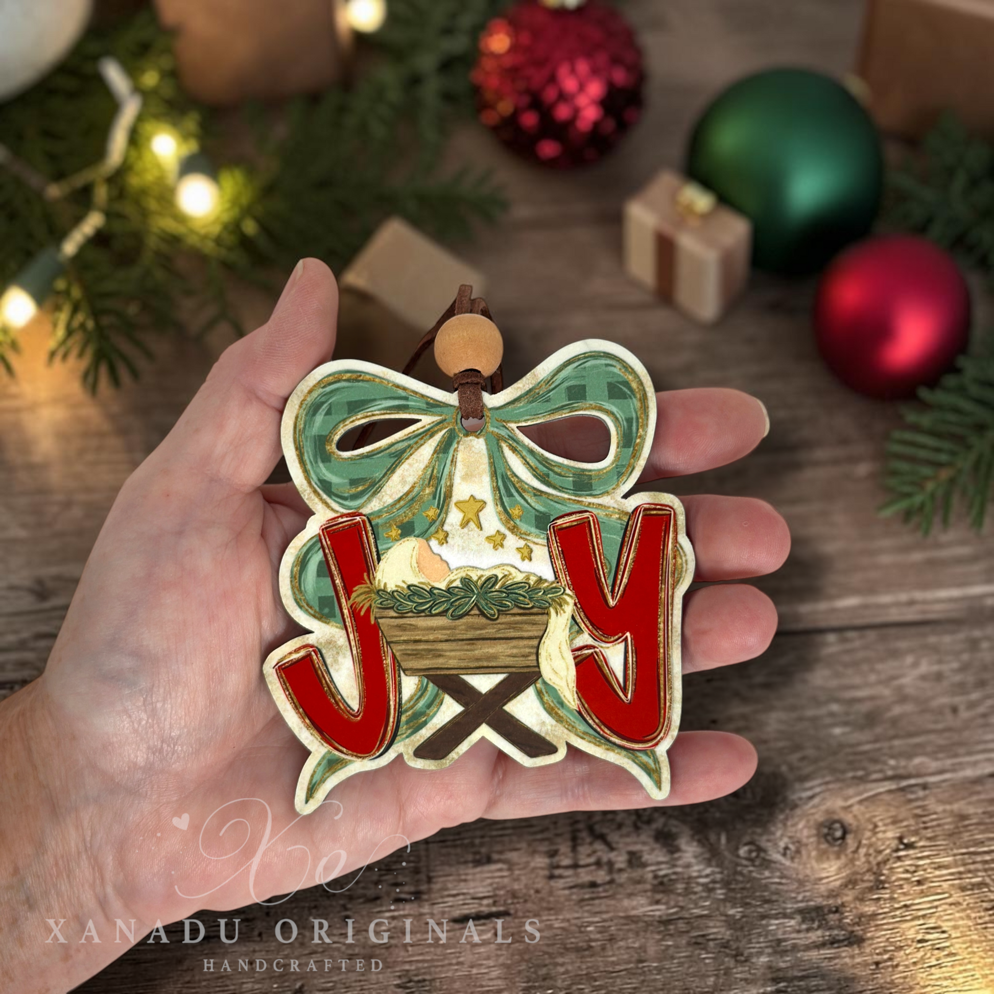 A hand holds a handcrafted Christmas ornament shaped like the letter J and Y with a nativity scene and a green bow. The background shows a rustic wooden table with pine branches, lights, and ornaments.